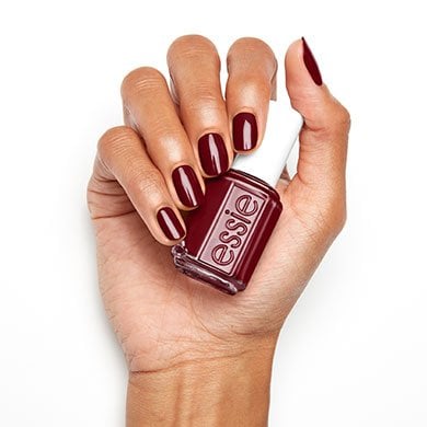 Hand holding a bottle of Essie nail polish, with fingers painted in a glossy, deep red shade.