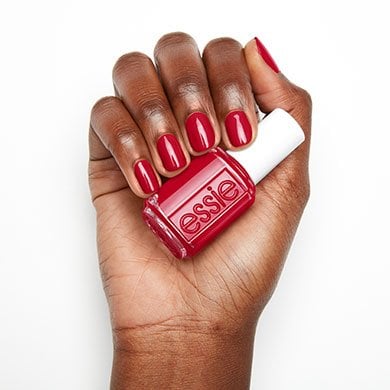 A hand with smooth, dark skin displays short, neatly manicured nails painted in vibrant red, holding a matching red nail polish bottle labeled "essie."