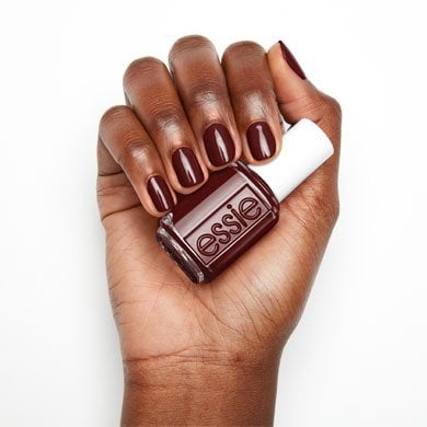 A hand holding a bottle of Essie nail polish in deep burgundy. The nails are painted the same color, conveying elegance and sophistication.