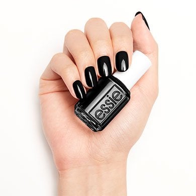Hand with glossy black nails holding a bottle of black Essie nail polish against a white background, conveying a chic and stylish tone.