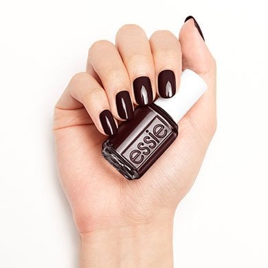 Hand with dark maroon nails holds an Essie nail polish bottle. The polished nails match the bottle color, set against a plain white background.
