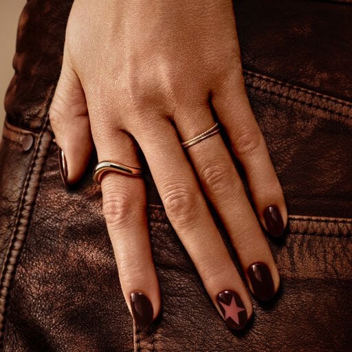 Hand with dark brown nail polish and a star design on one nail rests on brown leather. Gold rings on fingers.
