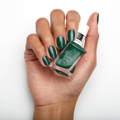 A hand with polished nails holds an Essie bottle. The nails are painted dark green, matching the polish. The background is plain white.