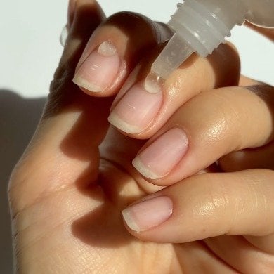 A hand with neatly trimmed, natural nails is applying a drop of cuticle oil from a small bottle.