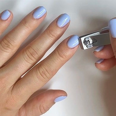 Light skin hands wearing lilac gel nails, one hand trimming a nail with clippers.