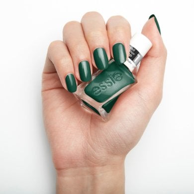 Hand with green nails holds an Essie nail polish bottle. The polished nails match the bottle color, set against a plain white background.