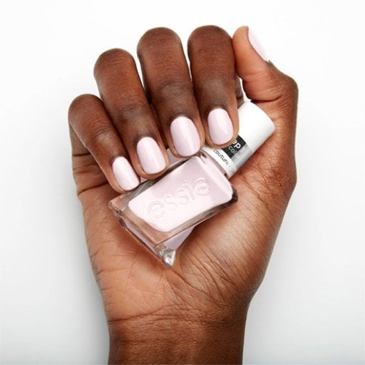 A hand with smooth, dark skin displays short, neatly manicured nails painted in soft pink, holding a matching pink nail polish bottle labeled "essie."