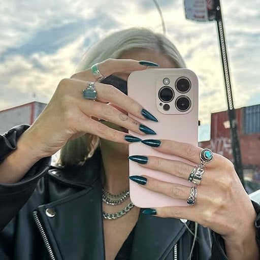 Model with dark blue manicure standing outside and holding phone in white phone case against a cloudy sky