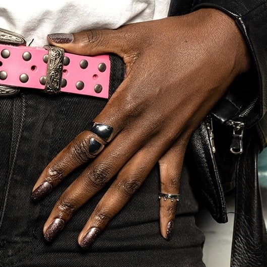 Model wearing black glitter nail polish and statement rings with hand placed over jean pocket