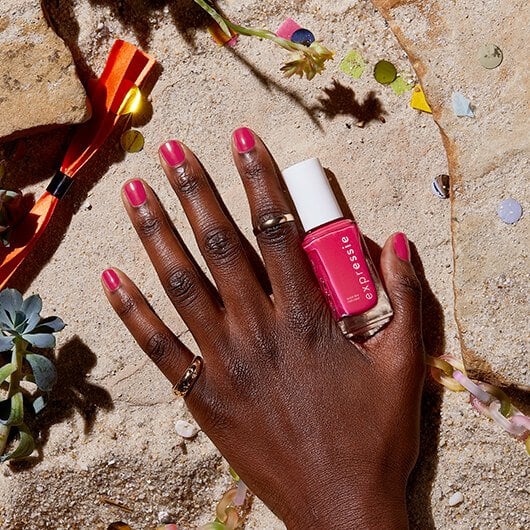 Model's pink manicured hand and bottle of pink nail polish placed against a rocky beach background